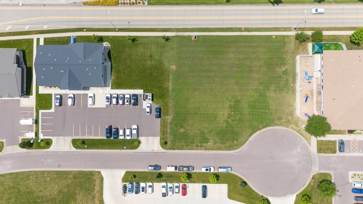 Aerial view of a tan industrial complex with flat white roofs, vast parking lots, and a dense green forest background.