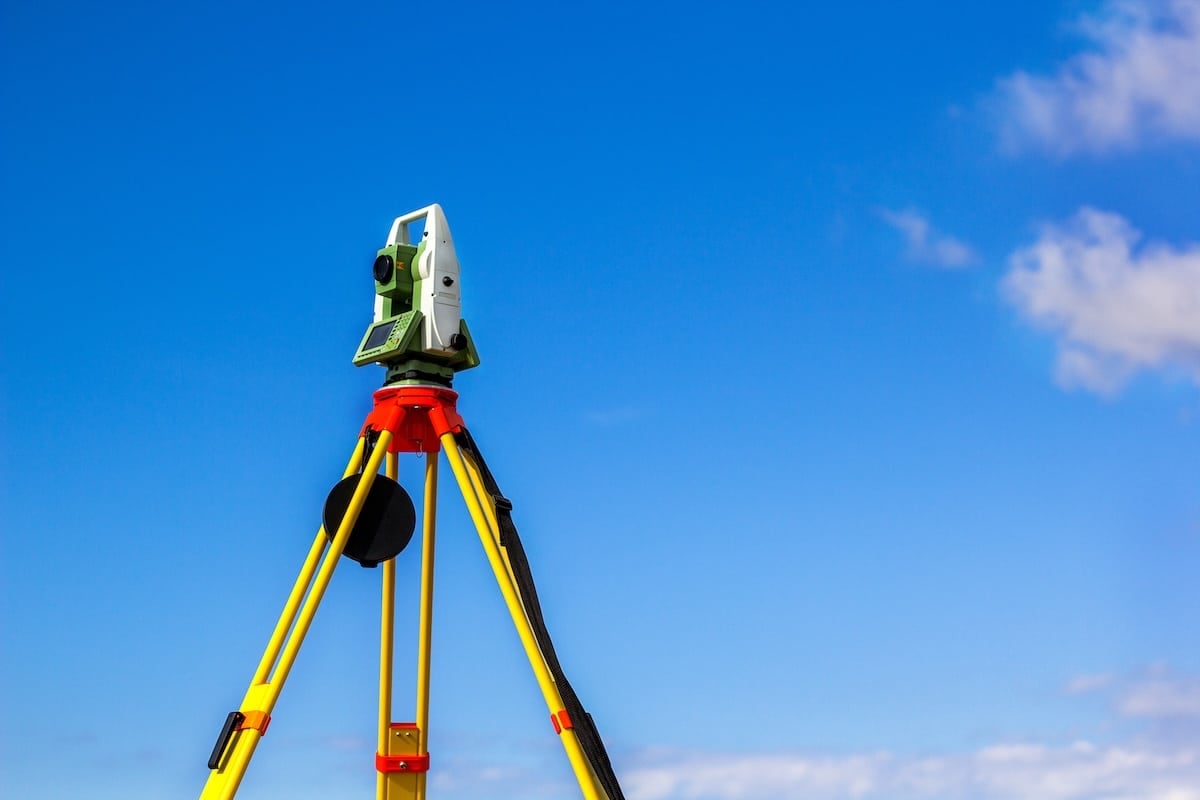 A white and green theodolite on a yellow tripod against a clear blue sky with light, wispy white clouds.