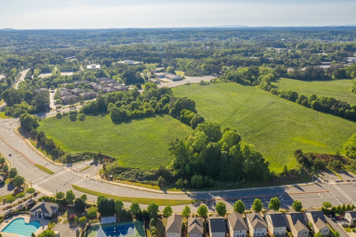 Aerial view of green fields, grey-roofed houses, a blue swimming pool, and a winding road through a dense treeline.