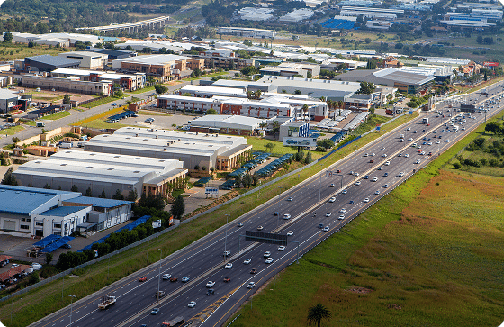 Diagonal highway traffic separates a green field from a dense industrial park with white flat-roofed buildings.