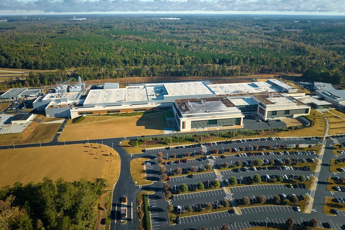 Aerial view of a tan industrial complex with flat white roofs, vast parking lots, and a dense green forest background.